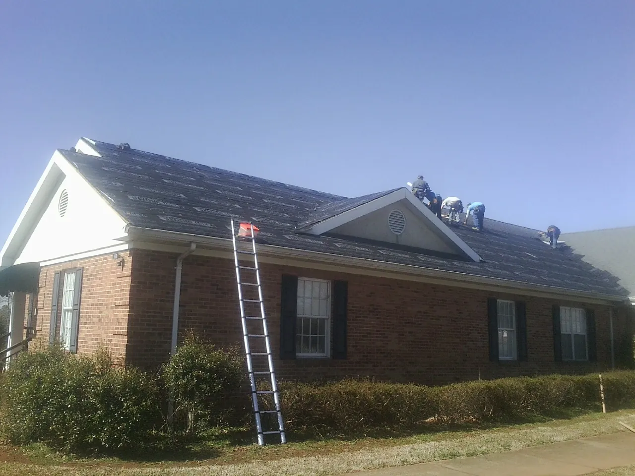 Crew tearing shingles off a brick ranch roof with a ladder set — Metal Roof Repair work by our crew in Osco