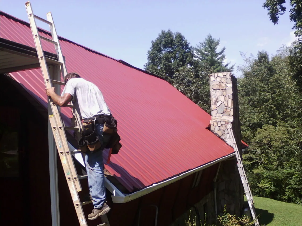 Roofer on a ladder beside a red metal roof and stone chimney — Soffit & Fascia Repair by Roofing Brothers in Osco
